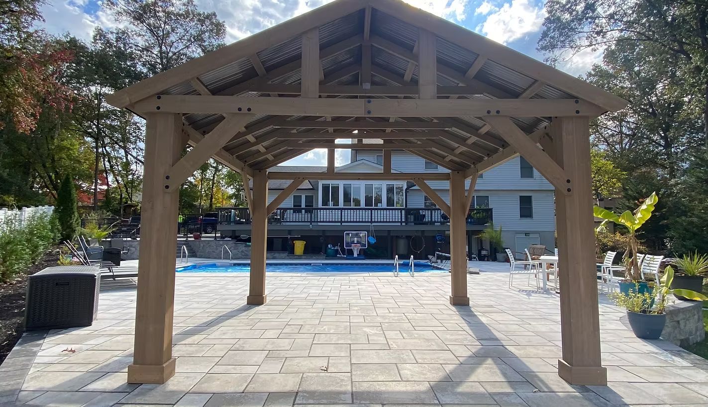 Wooden pavilion over a stone patio beside a pool, with a building and trees in the background
