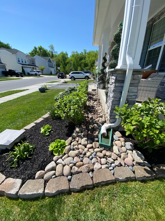 Front yard garden bed with river rocks, shrubs, and downspout beside a house on a suburban street