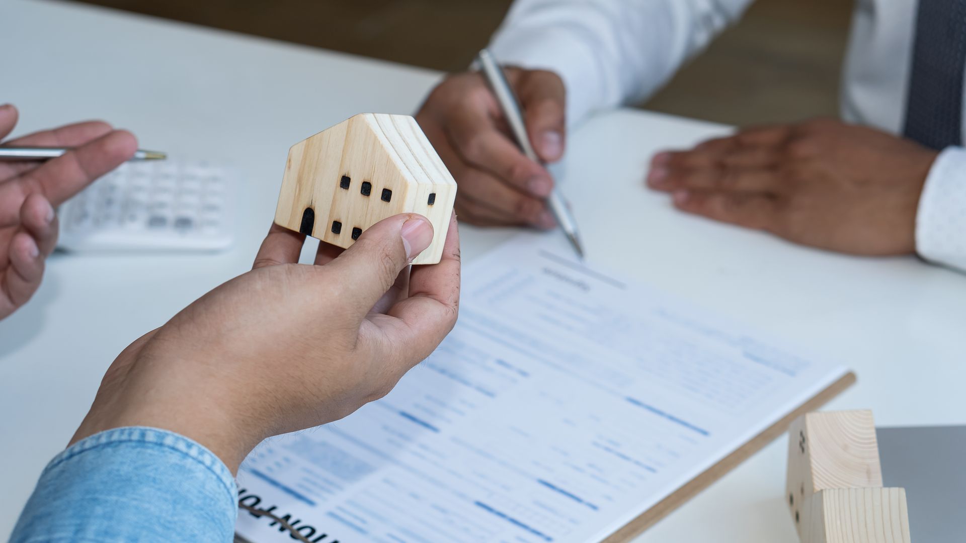 Hands holding a wooden house model over a contract; two people discuss and sign documents at a desk.
