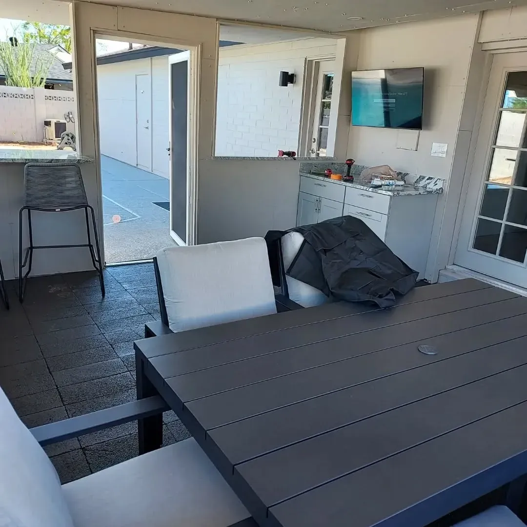 Patio with a gray table and white chairs. TV mounted on the wall, outdoor setting.