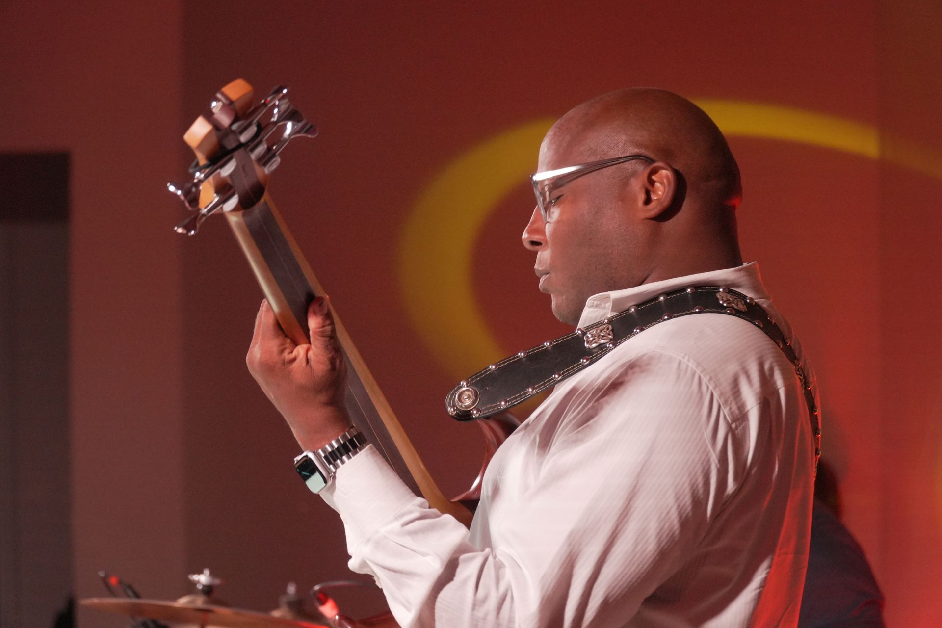 Bald Black man playing bass guitar, wearing glasses, in a warmly lit venue.