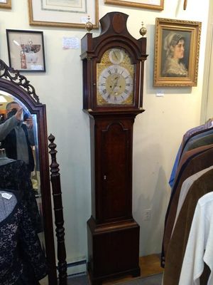 A tall, dark wood grandfather clock stands against a wall, surrounded by framed art and a mirror in a store.