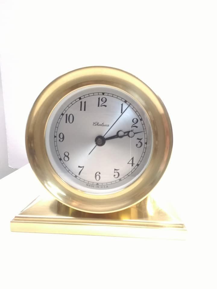 A brass Chelsea desk clock with a circular silver face, Roman numerals, and a polished metallic base on a white surface.