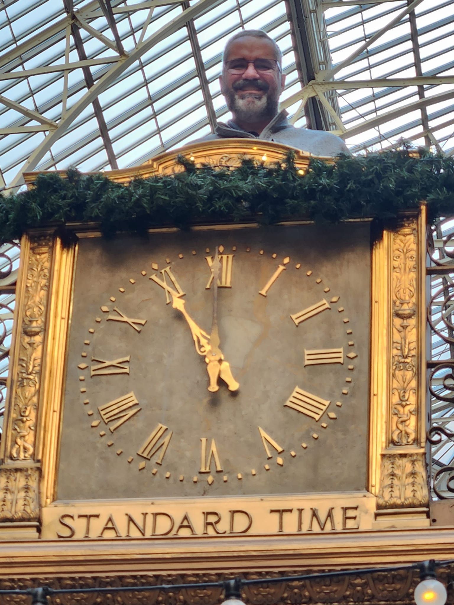 A smiling person stands behind an ornate, gold-framed clock labeled 