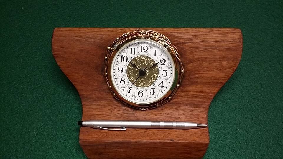 A gold-rimmed analog clock is mounted on a wooden stand with a silver pen resting in a slot below it on a green surface.