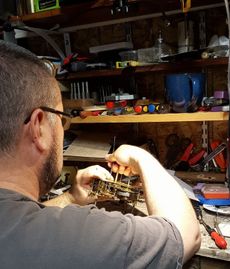 A person wearing glasses works on a small mechanical device at a workbench cluttered with tools and supplies.