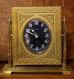 A vintage gold-toned mantle clock with a black oval face, ornate frame, and vertical side supports on a rectangular base.