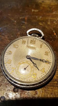 A close-up of a round, vintage silver pocket watch with gold-colored numerals on a textured, light-colored dial.