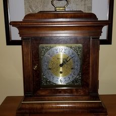 A wooden mantle clock with a metallic face, gold-tone center, and black numerals, sitting on a table.