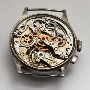 An open-cased, silver-toned mechanical chronograph watch movement with exposed gears, levers, and balance wheel.
