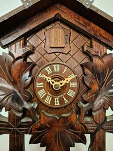 Close-up of a carved wooden cuckoo clock face with Roman numerals, ornate gold hands, and leaf detailing.