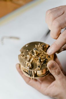 Hands work carefully on a golden, disassembled mechanical watch movement on a white surface.