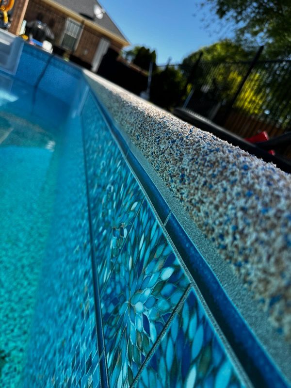 Close-up of a pool's edge: speckled coping, patterned blue tile, and clear water on a sunny day.