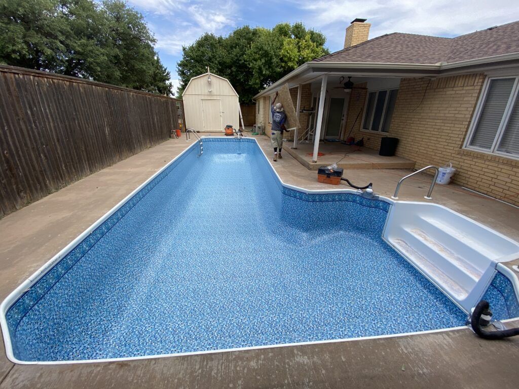 Rectangular in-ground swimming pool with blue mosaic-patterned liner, concrete surround, and a person standing near the house.
