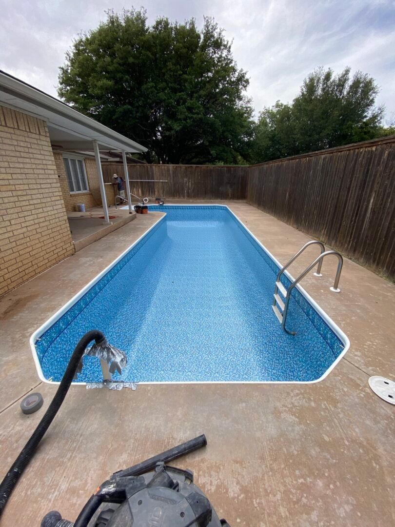 Rectangular, blue-tiled swimming pool surrounded by concrete, near a house with a fence and trees.