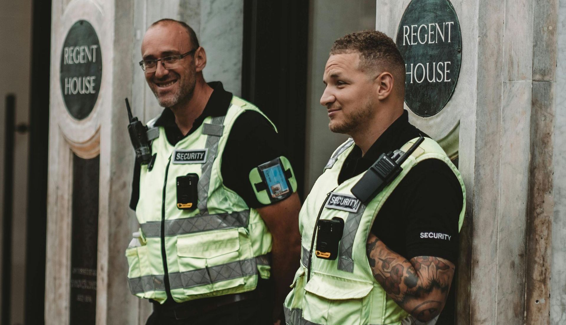 Two security guards in reflective vests stand outside a building. One smiles, the other has crossed arms.