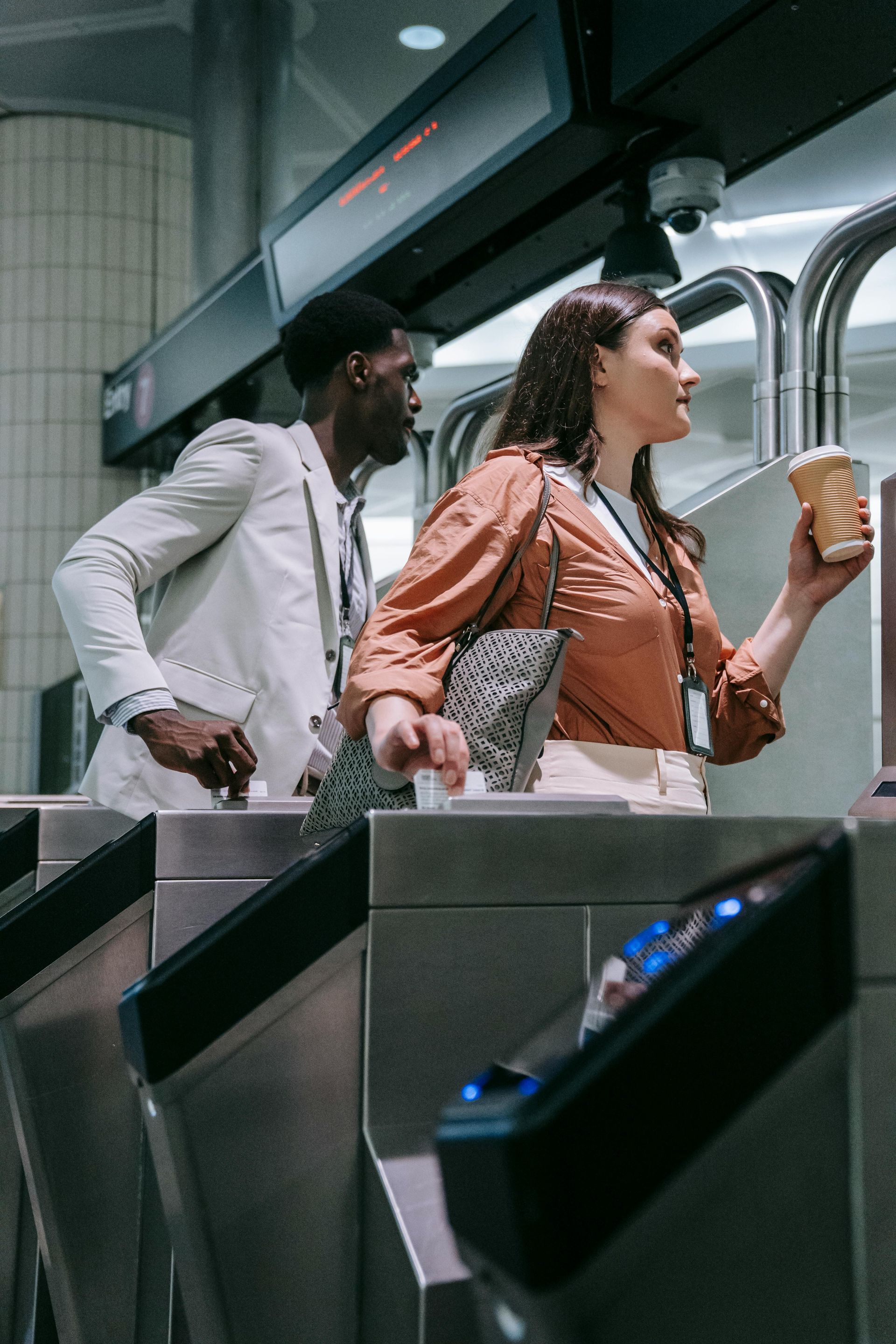 People at subway turnstile: woman holding coffee, man reaching, both rushing past metal gates.