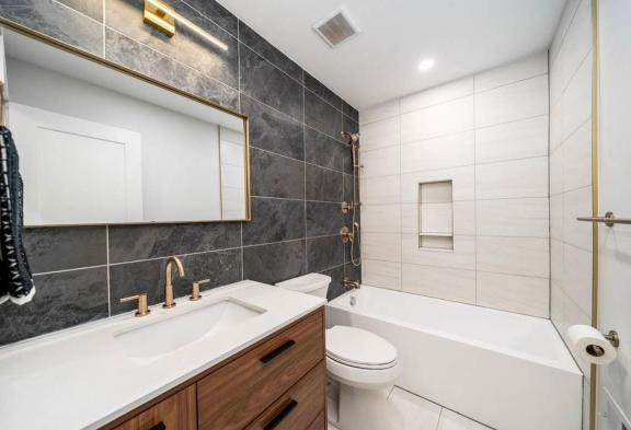 Modern bathroom with dark gray accent wall, gold fixtures, wood vanity, and white tile.