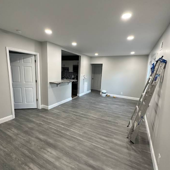 Empty, newly-painted living room with gray flooring. A ladder leans against the wall. Kitchen visible in the background.