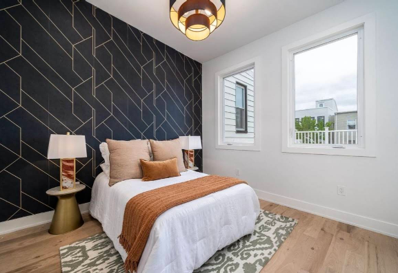 Bedroom with white walls, patterned accent wall, bed with throw, windows, rug, and bedside lamps.