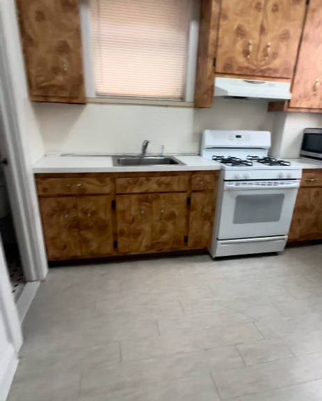 Kitchen with wood cabinets, white stove, sink, and countertops. Tan floor, window with blinds.