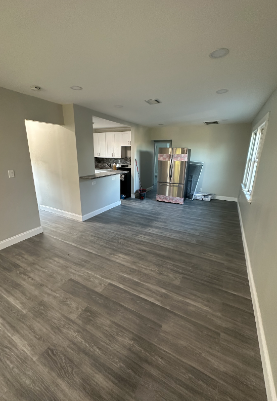 Empty living room with wood-look flooring, light gray walls, and an opening to a kitchen.