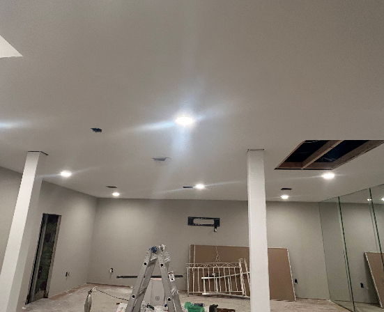 Interior room under construction with white walls, ceiling, and support beams; recessed lights and exposed opening in ceiling.
