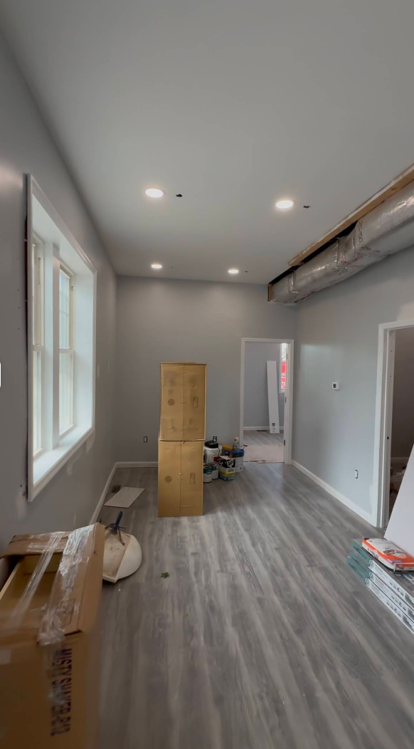 Empty room under construction with gray walls and wood flooring, with a window and cardboard boxes present.