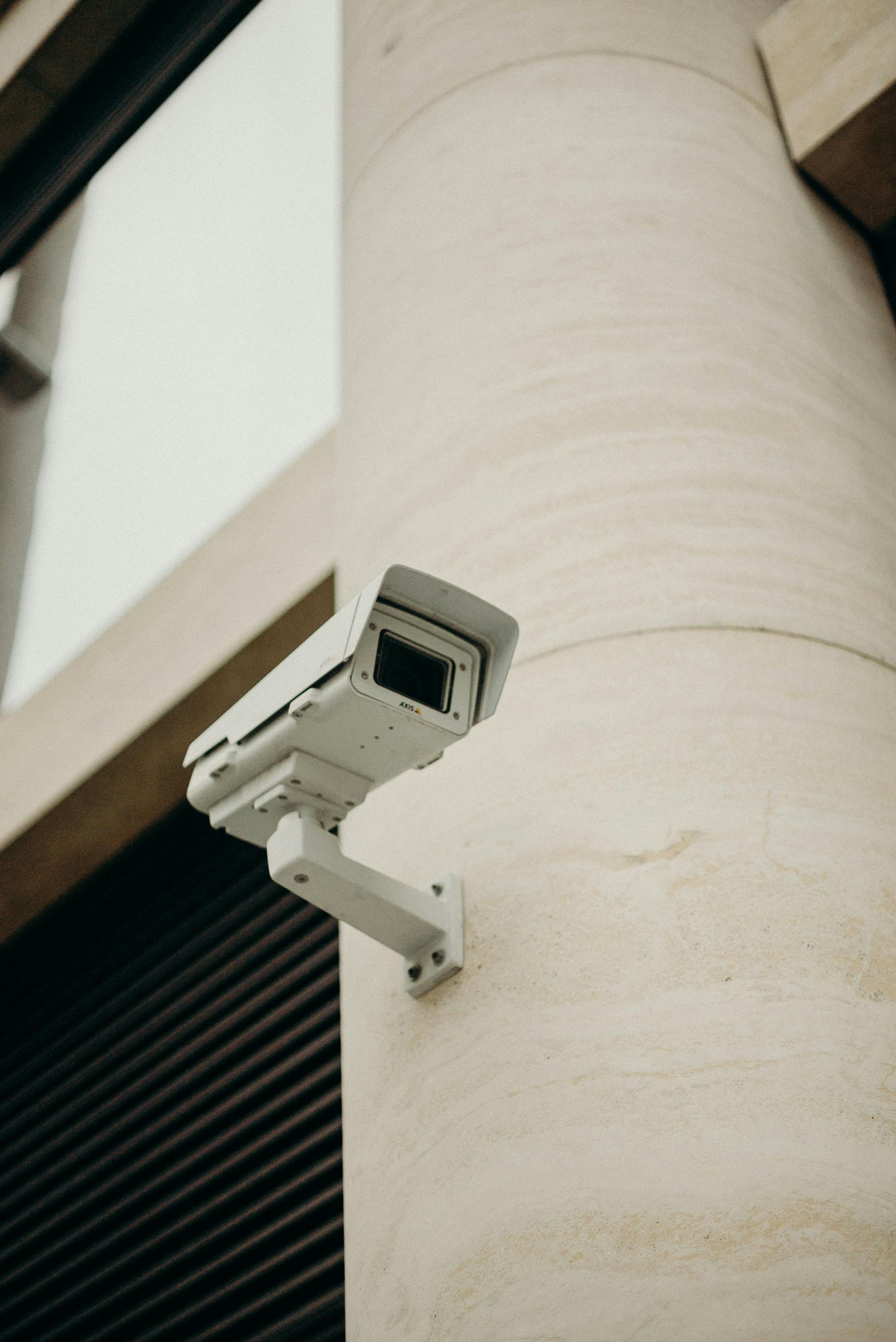 Una telecamera di sicurezza di colore chiaro montata su un pilastro cilindrico beige accanto alla facciata esterna di un edificio con rivestimento a listelli scuri.