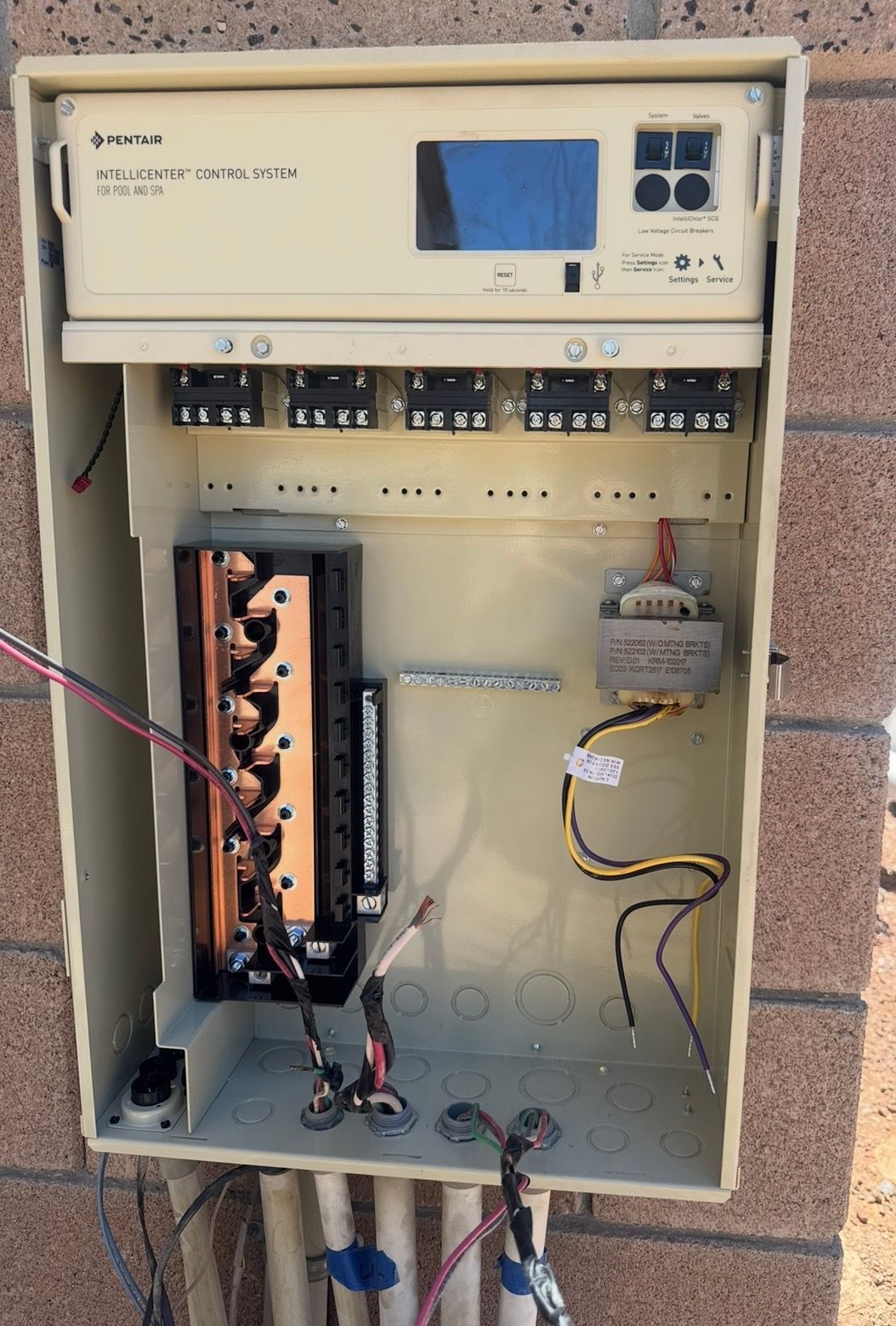 Open electrical box mounted on a brick wall. Beige components, wiring, and a digital display are visible inside.