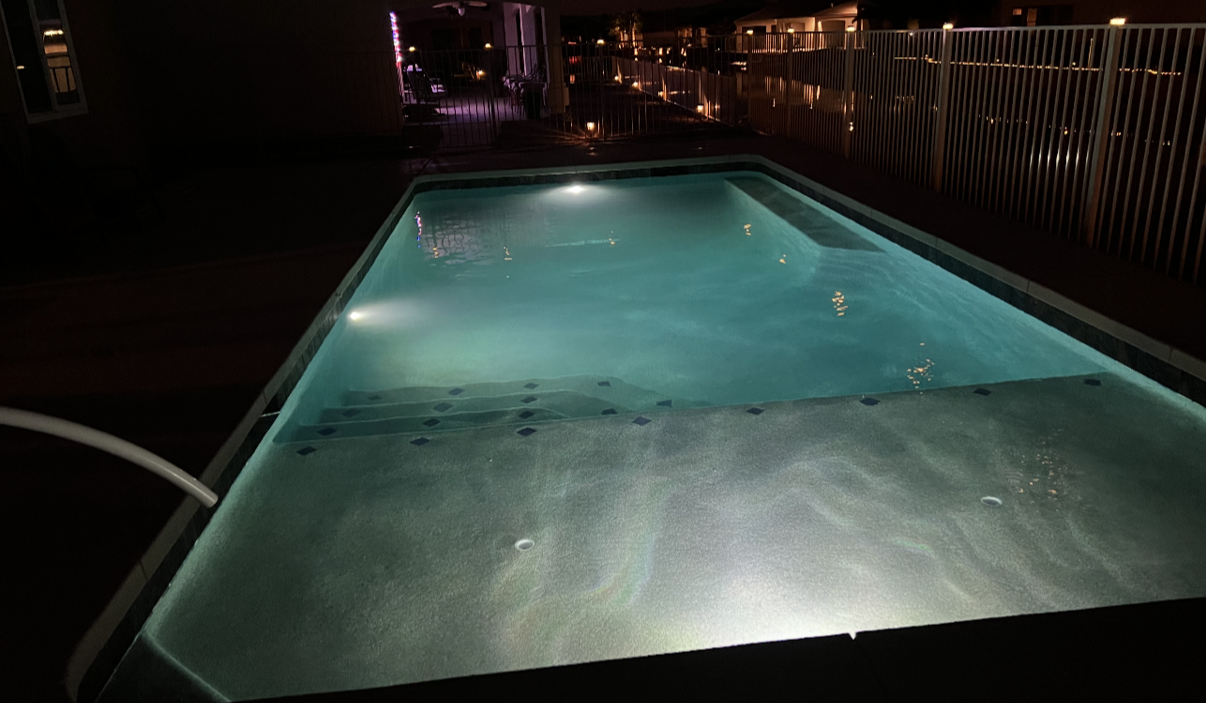 Nighttime view of a rectangular pool with illuminated water; metal fence and buildings in the background.