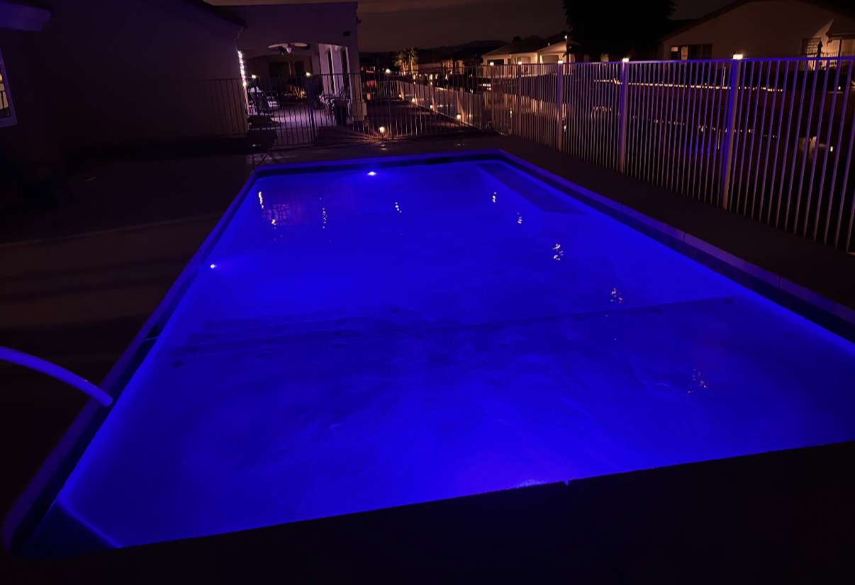 Pool at night with blue lights; fence on the right, deck around the pool, and dark sky.