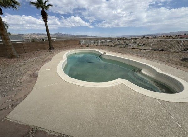 An empty kidney-shaped pool with a concrete deck. Palm trees and a desert landscape in the background.
