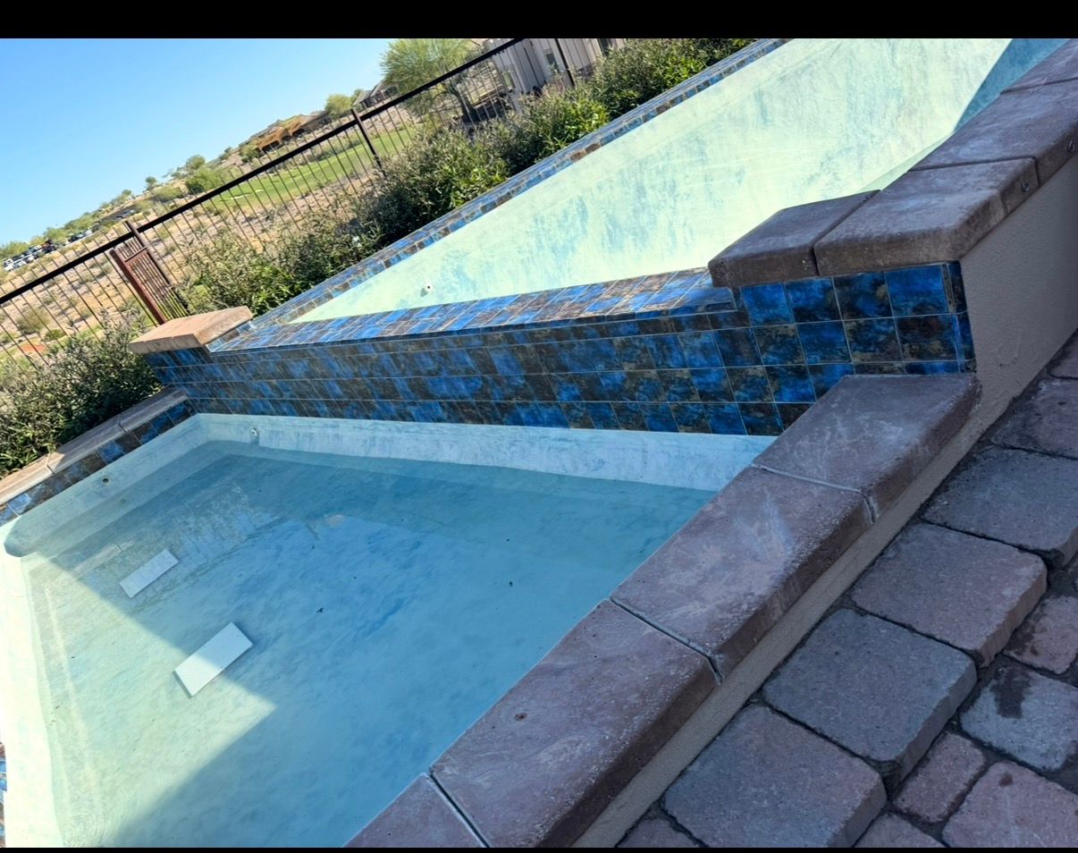 Empty rectangular swimming pool with blue-tiled wall feature, grey stone coping, and dry desert backdrop.
