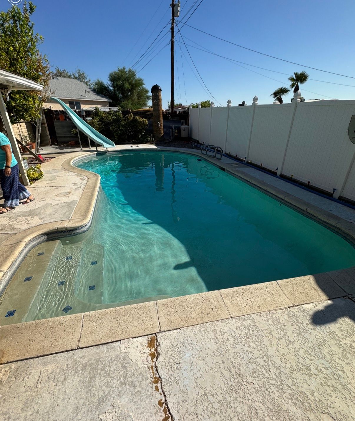 A backyard pool with a slide and white fence, under a clear, sunny sky.