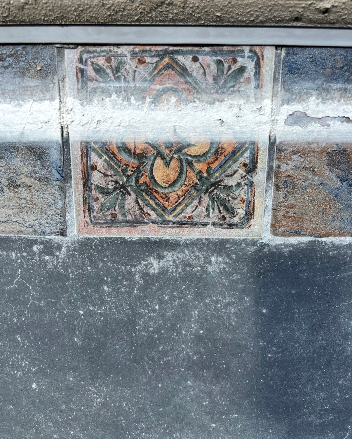 Ornate, faded tiles with floral designs, framed above a dark concrete surface.