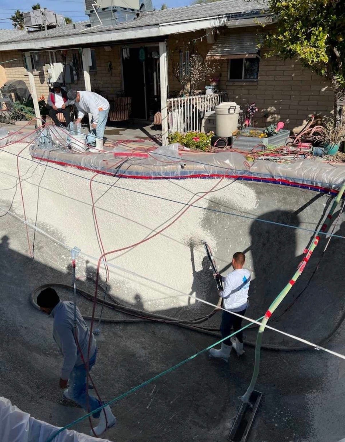 Workers resurfacing a swimming pool. Three people in the pool, house in the background, cables and hoses throughout.