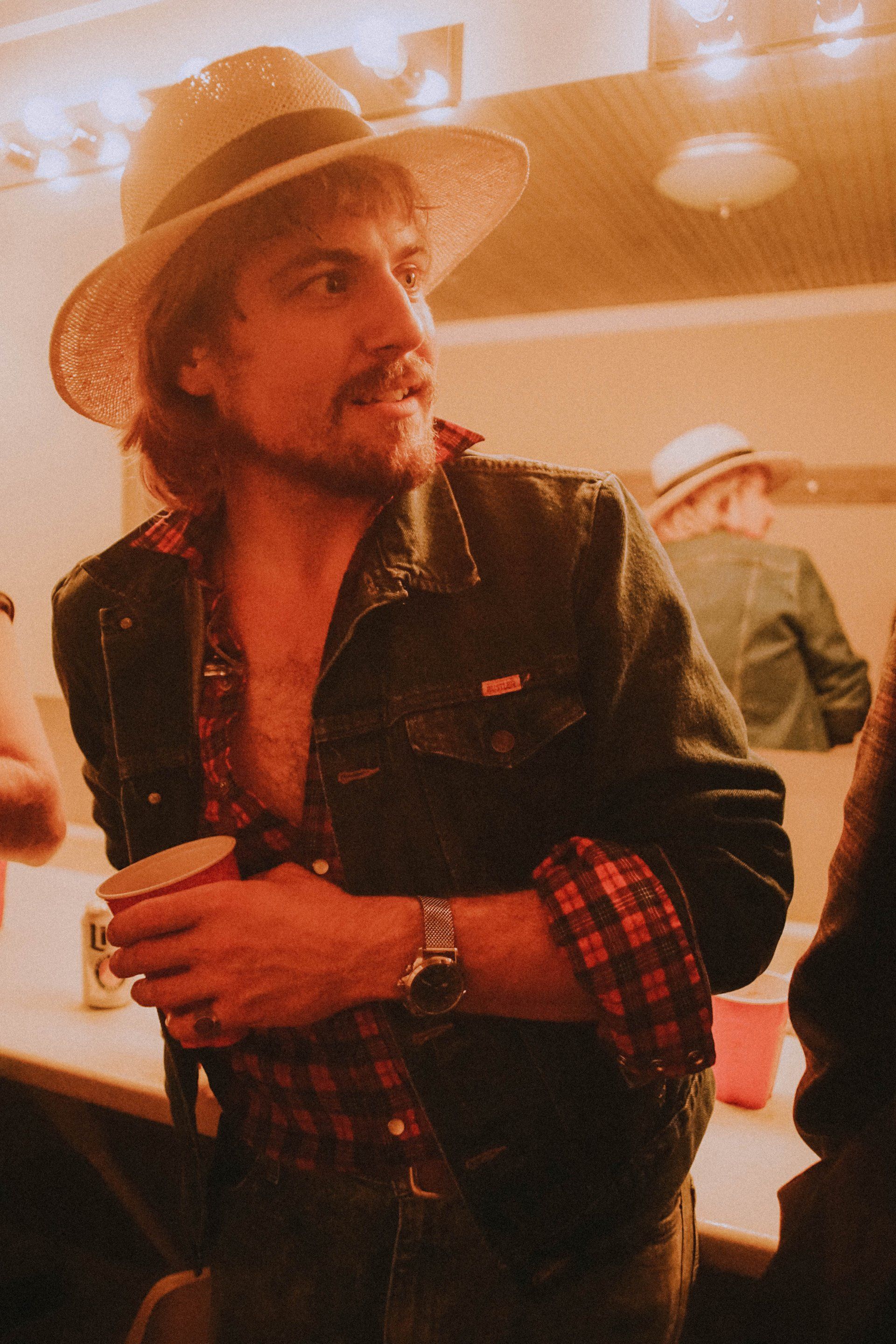A man wearing a hat and a plaid shirt is standing in front of a mirror holding a cup.