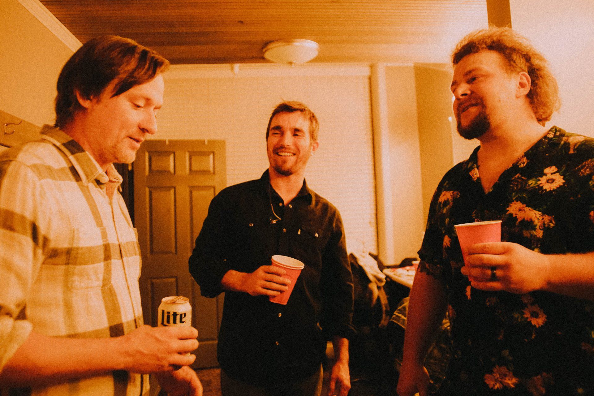 Three men are standing in a room and one of them is holding a can of lite beer