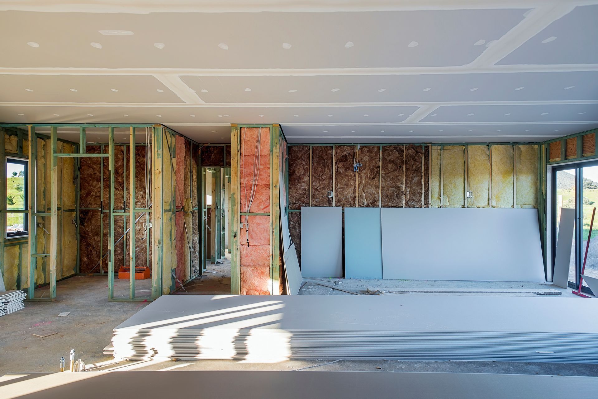 Interior under renovation with exposed framing, insulation, and drywall stacked against a partially finished wall.