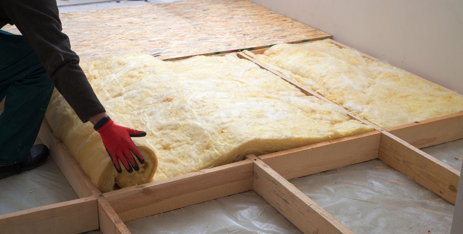 Person in red gloves installing yellow insulation between wooden floor joists