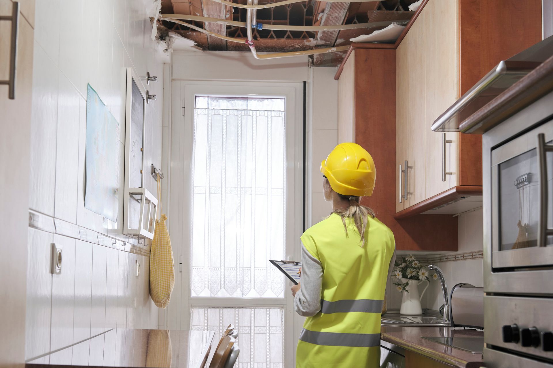 Worker in a yellow hard hat inspecting exposed ceiling wiring in a narrow kitchen.