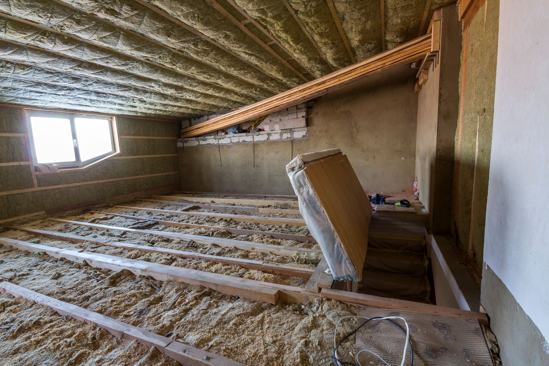 Insulated attic under construction with exposed framing, a window, and a mattress leaning against the wall
