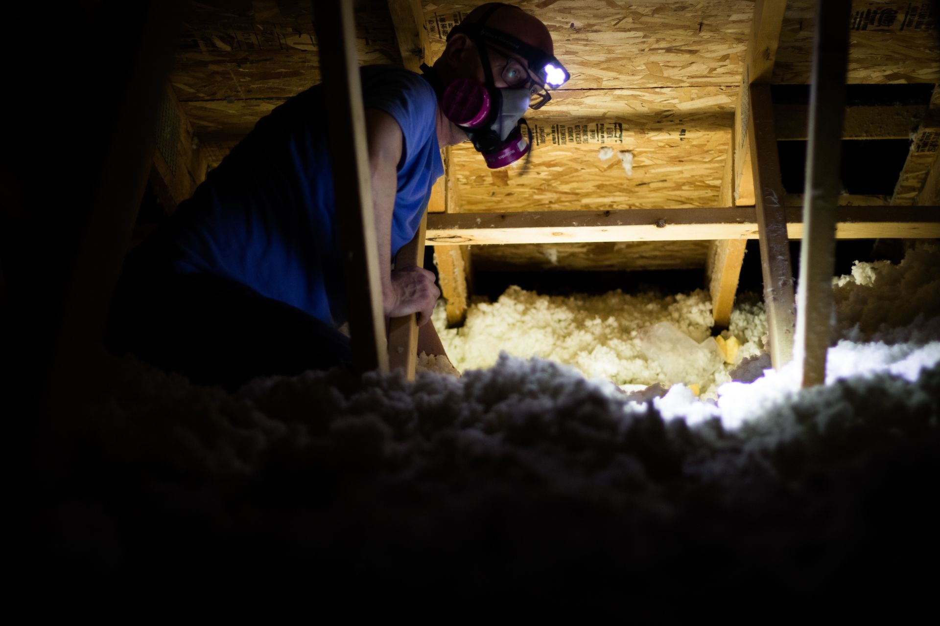 Person in a headlamp and mask inspecting insulation in a crawl space under a house