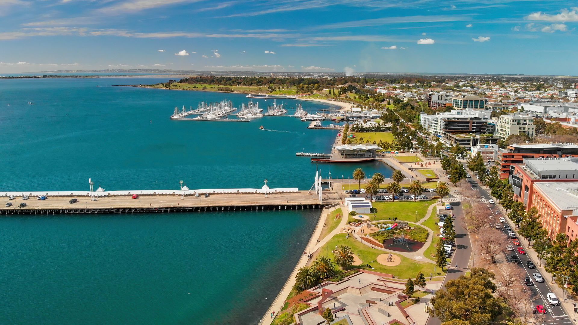 Aerial view of a turquoise harbor, long pier, and coastal city with roads and buildings under a blue sky