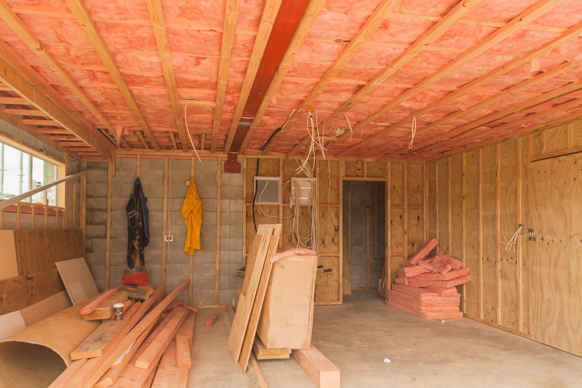 Unfinished room with exposed wooden framing, stacked lumber, and hanging work clothes.