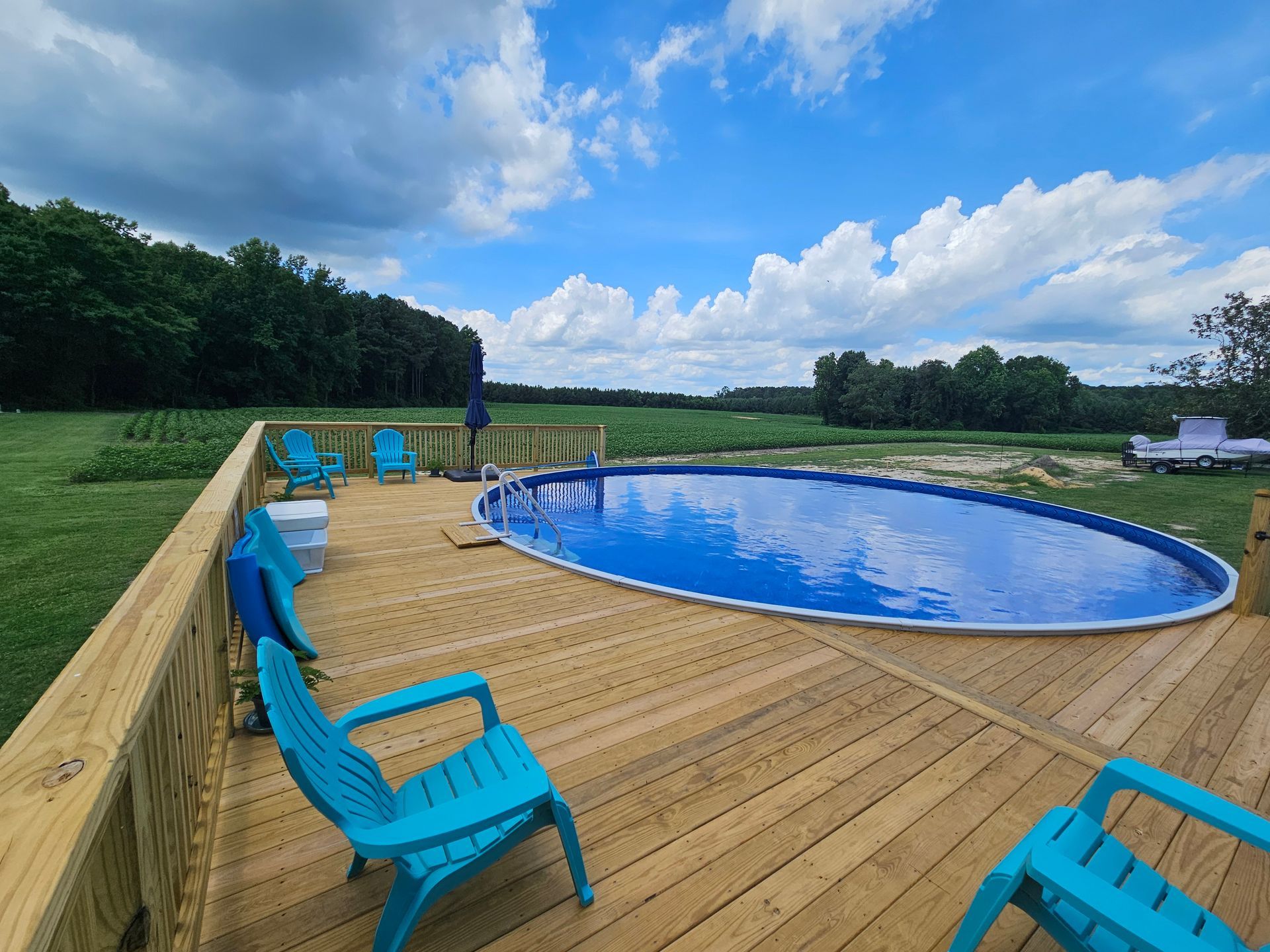 Curved blue pool on a wooden deck with turquoise chairs, overlooking open green fields under a cloudy sky