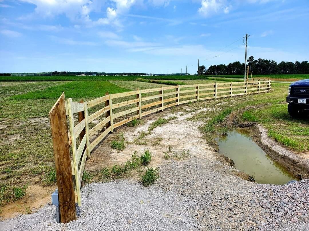 Curving wooden fence beside a gravel road and muddy puddles in a rural field under a bright sky