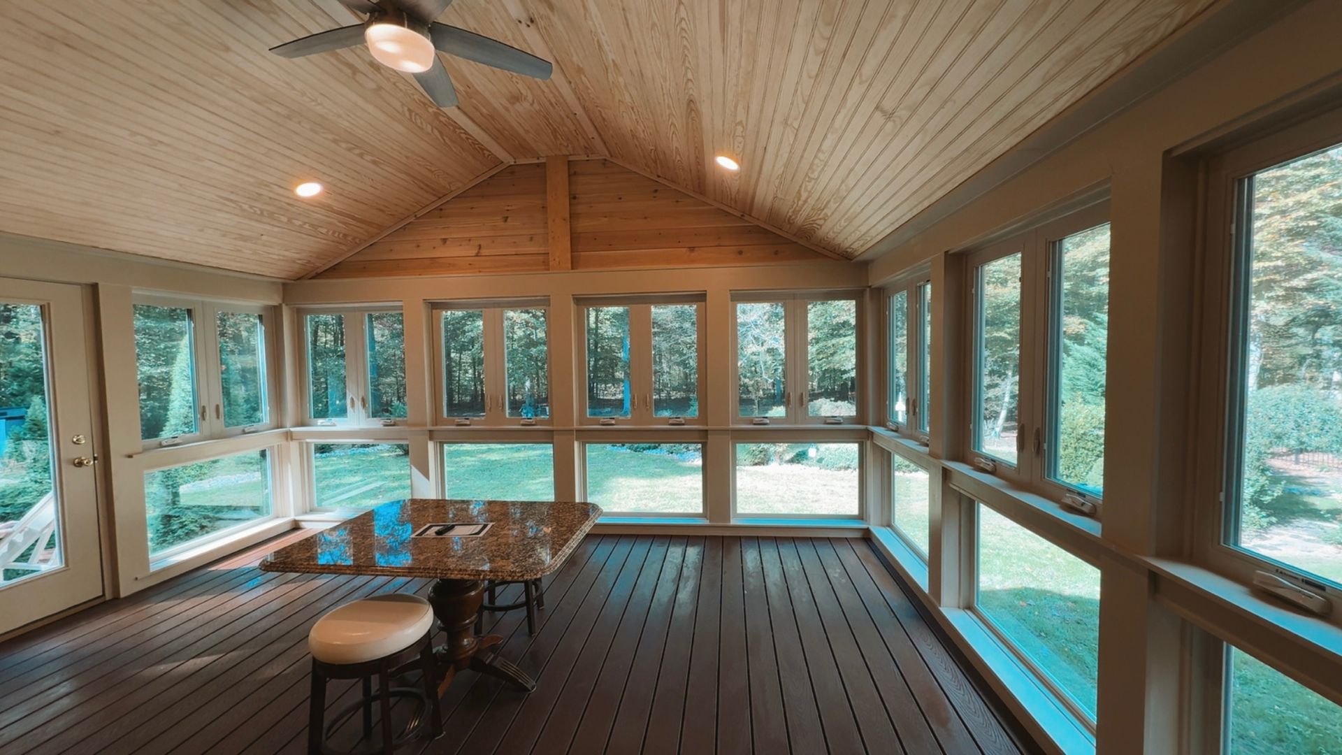 Sunroom with wooden ceiling, glass walls, and a table beside rows of empty chairs.