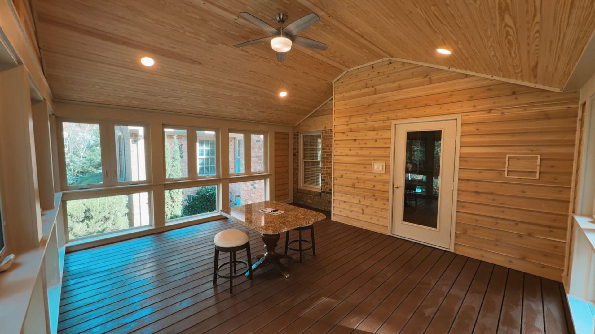 Wood-paneled sunroom with large windows, ceiling fan, and a table with chairs on a dark floor.